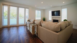 A spacious living room with a large beige sectional sofa, a flat-screen TV mounted above a fireplace, and a sliding glass door leading to a balcony. - Beverly Hills Luxury Condo, 5% Discounts on monthly bookings. (Los Angeles)
