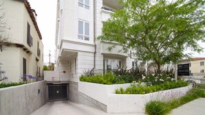 A modern building with a gated entrance, a landscaped area with plants and flowers, and a sign indicating a property for sale. - Beverly Hills Luxury Condo, 5% Discounts on monthly bookings. (Los Angeles)