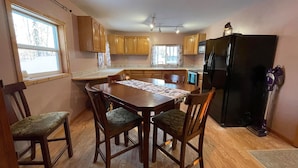 A kitchen with wooden cabinets, a dining table with chairs, a refrigerator, and a window with curtains. - Willow Forest Cabin with Sauna, Pet and Family Friendly (Willow)