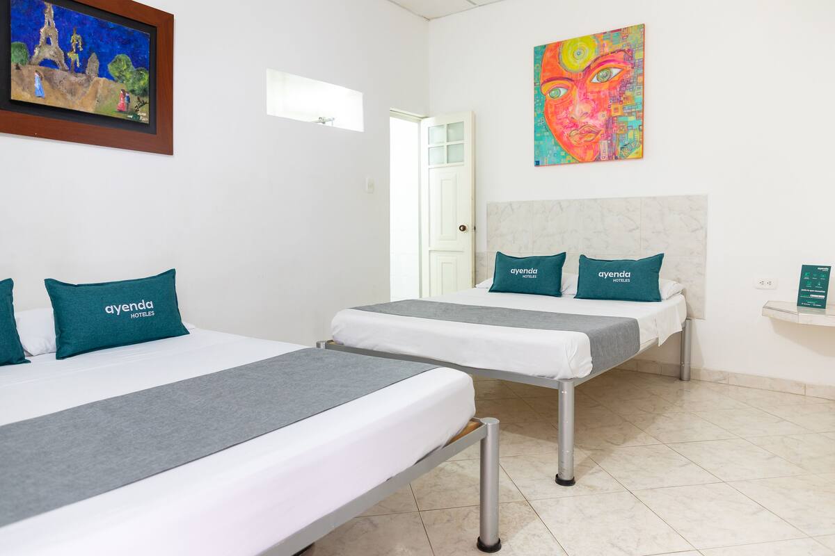 Two twin beds with grey and white bedding, teal pillows with 'ayenda HOTELES' text, a colorful abstract painting, and a marble headboard.