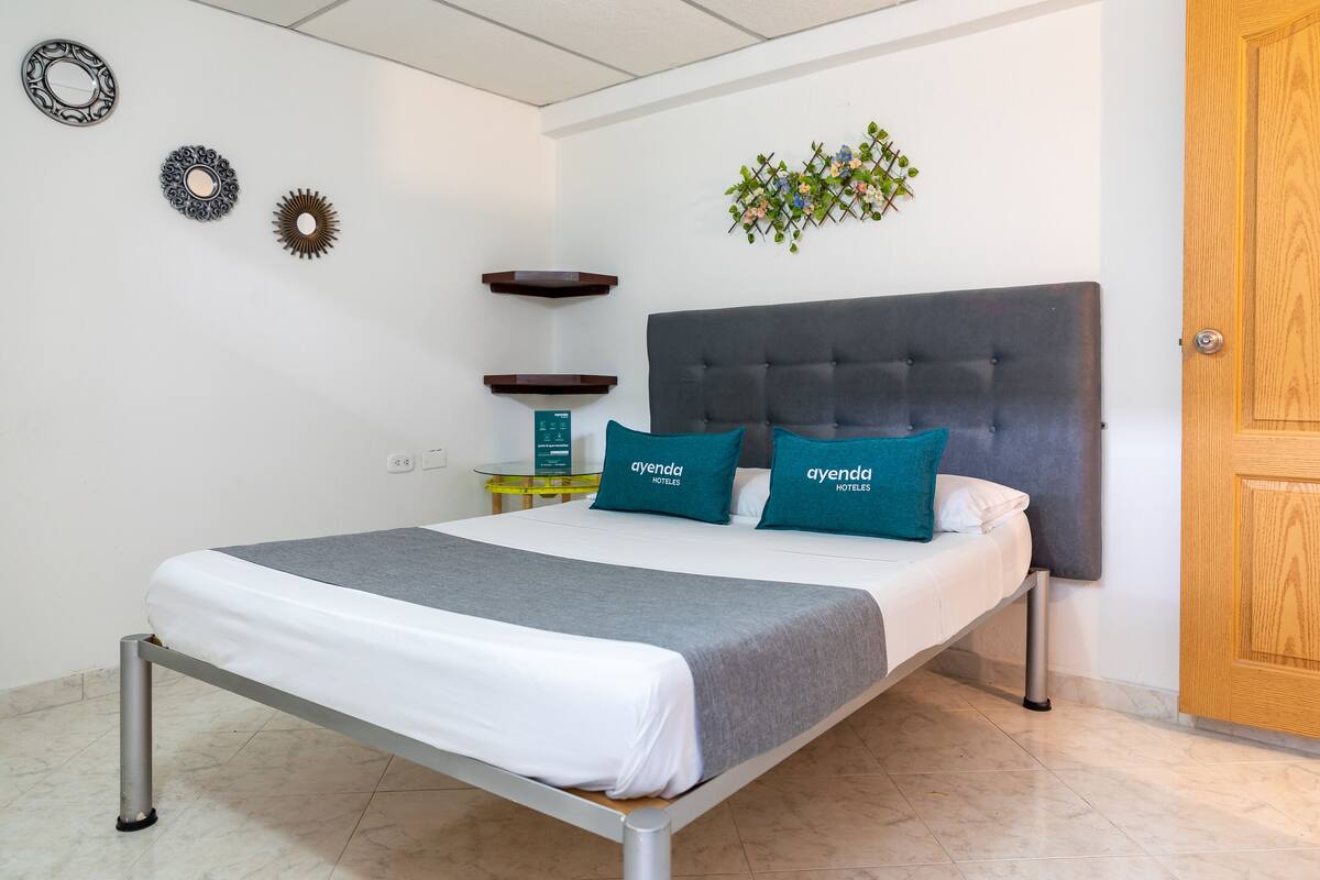 A neatly made bed with a grey and white bedding set, two teal pillows with "ayenda hotels" text, a grey headboard, and a wooden door to the right.