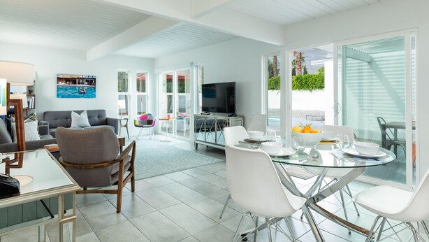 A modern living room with a glass dining table, white chairs, a grey sofa, and a flat-screen TV. - Racquet Club Escape (Palm Springs)