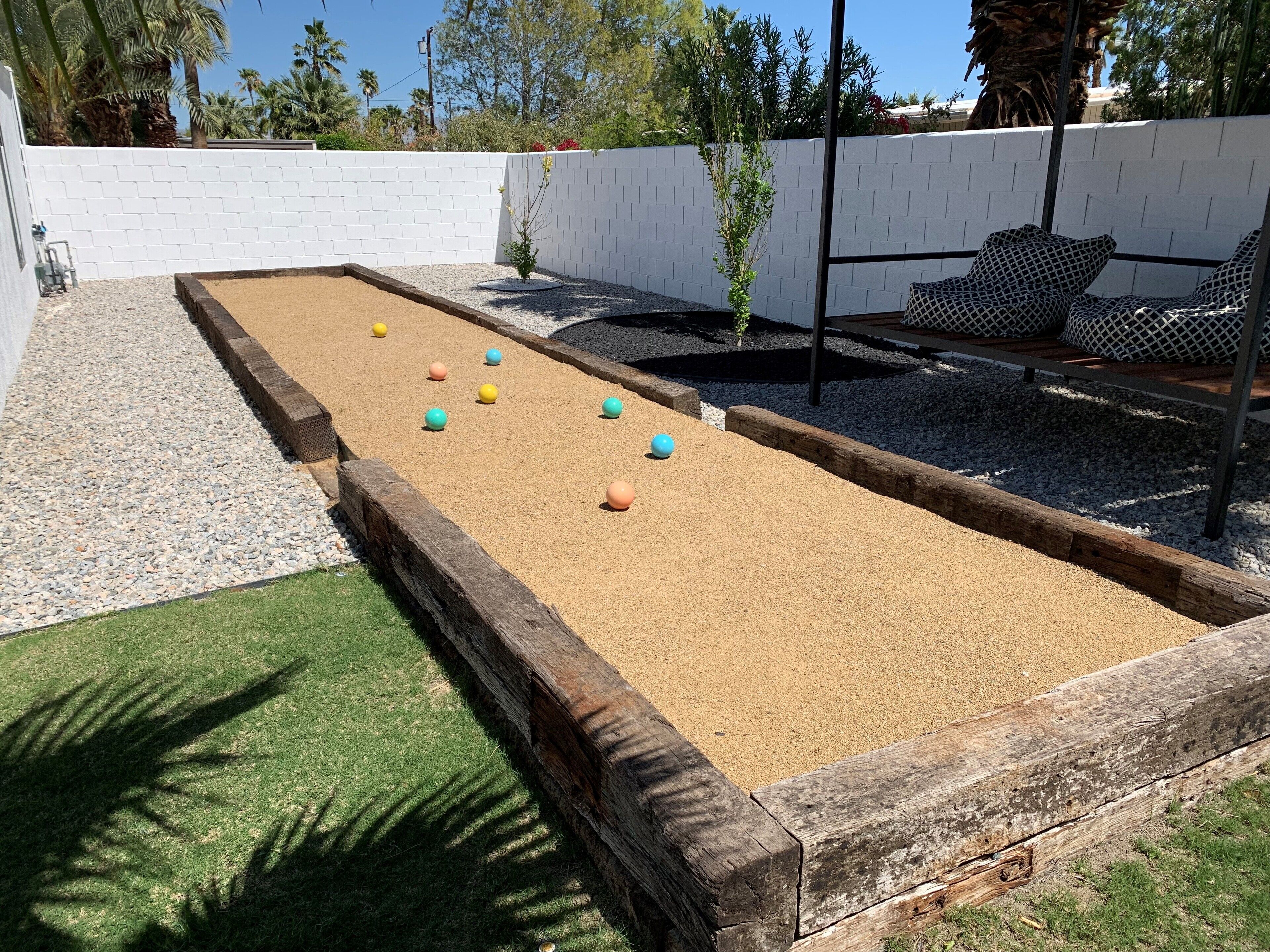 A croquet court with a wooden border, a white brick wall, and a garden area with a lawn and trees.