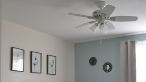 Une chambre avec un lit blanc, un ventilateur de plafond, un mur d’accent bleu et des œuvres d’art encadrées.