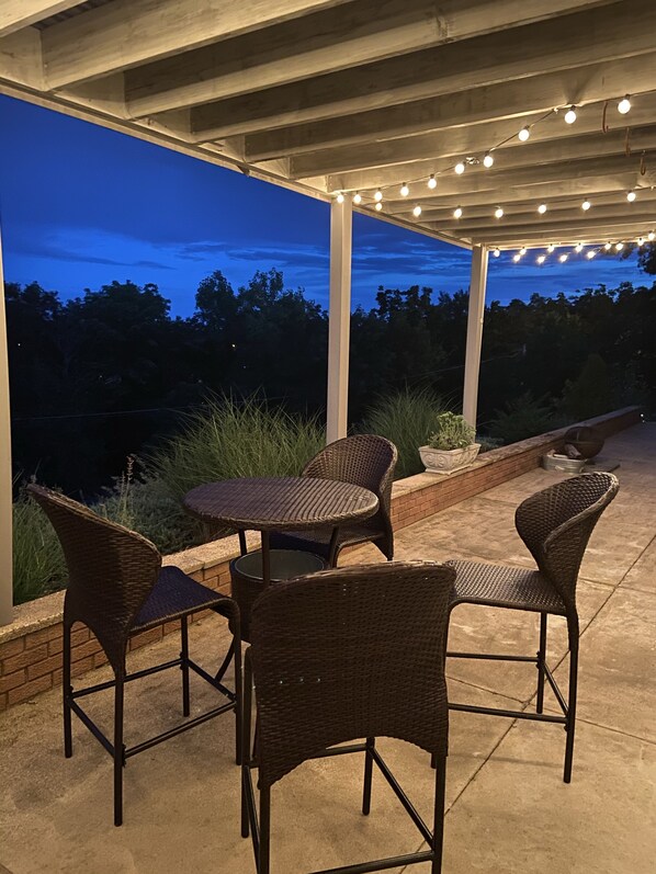 Une terrasse extérieure aménagée avec des meubles en osier, des guirlandes lumineuses et une vue sur les arbres au crépuscule.