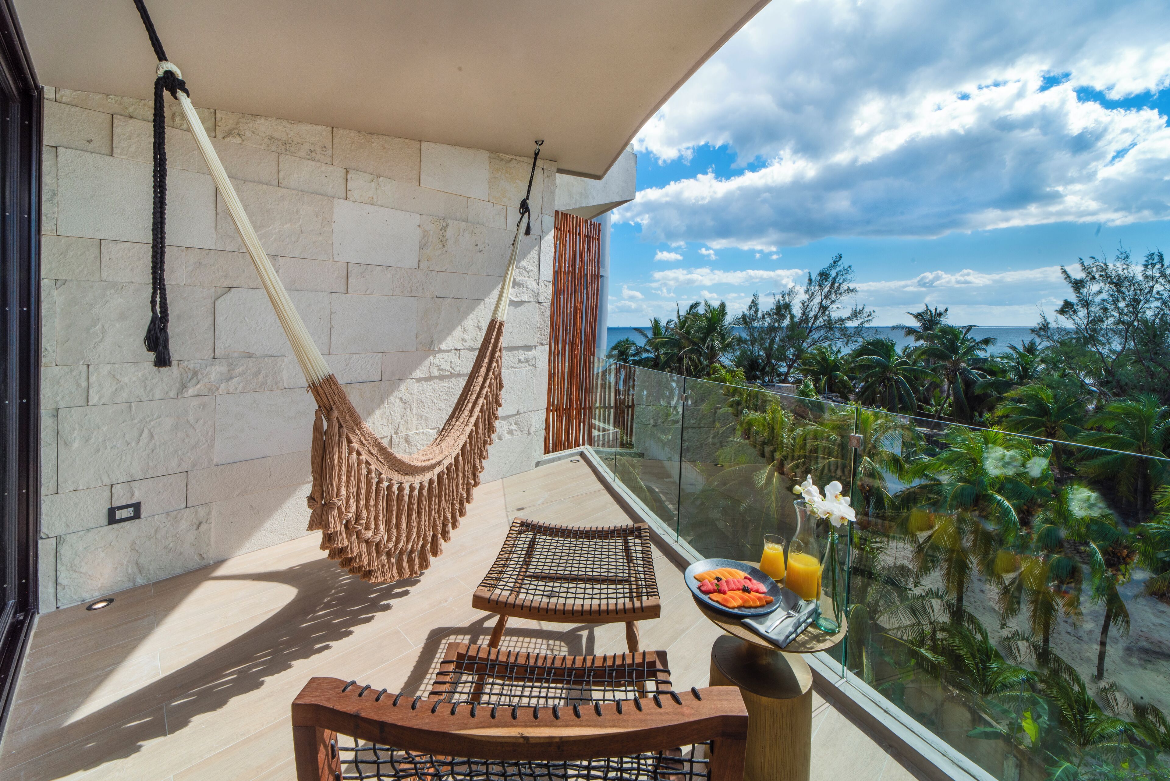 A balcony with a hammock, two wooden chairs, a glass table with a fruit plate and a vase of flowers, and a view of palm trees and the ocean.