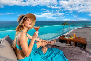 A person relaxing by a pool with a view of the ocean, a wooden table with drinks, and lounge chairs. - Mvngata Beach Hotel (Playa del Carmen)