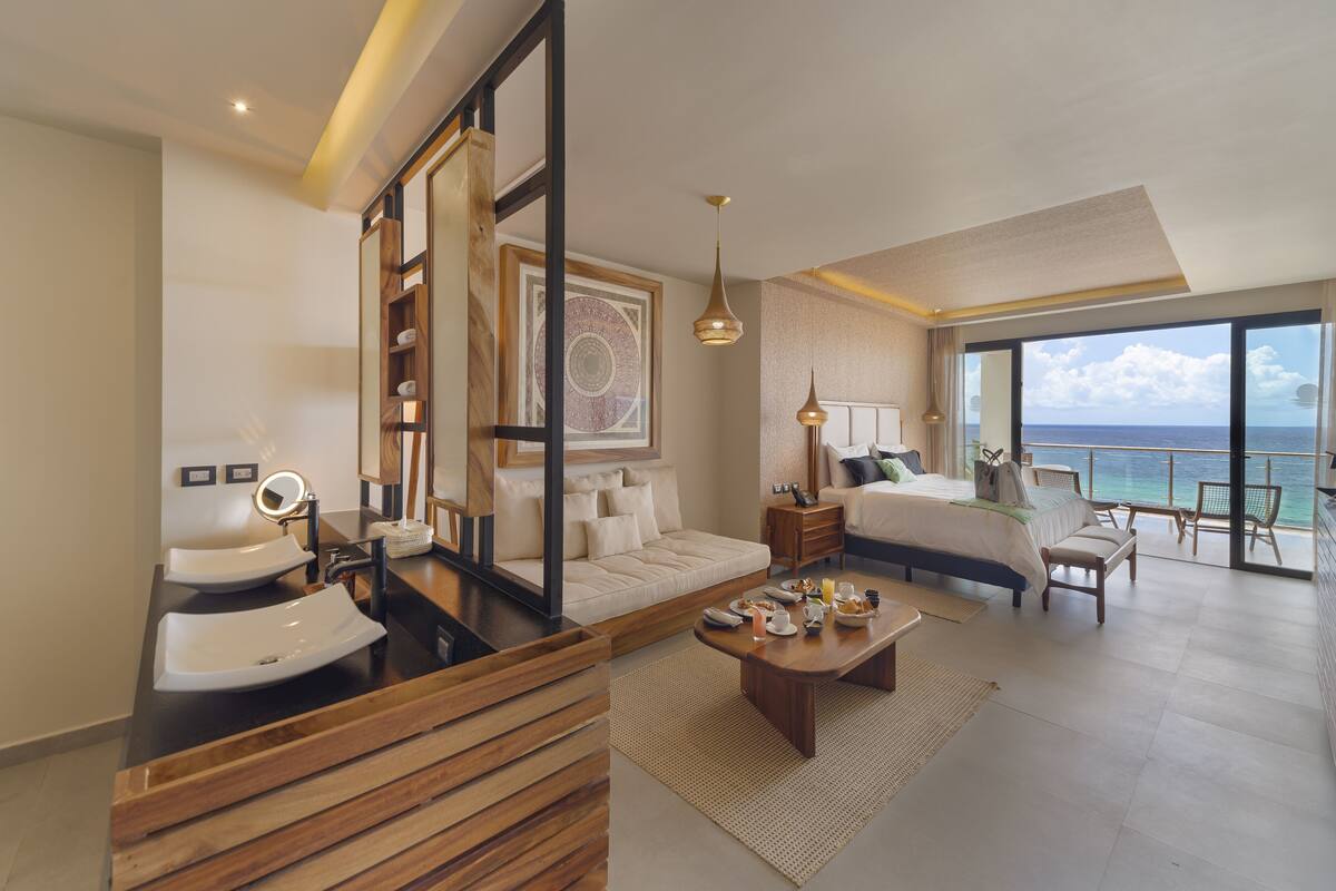 Une chambre d’hôtel moderne dotée d’un grand lit, d’un coin salon avec une table basse, d’une salle de bain avec un meuble-lavabo et un lavabo, et d’un balcon offrant une vue sur l’océan.