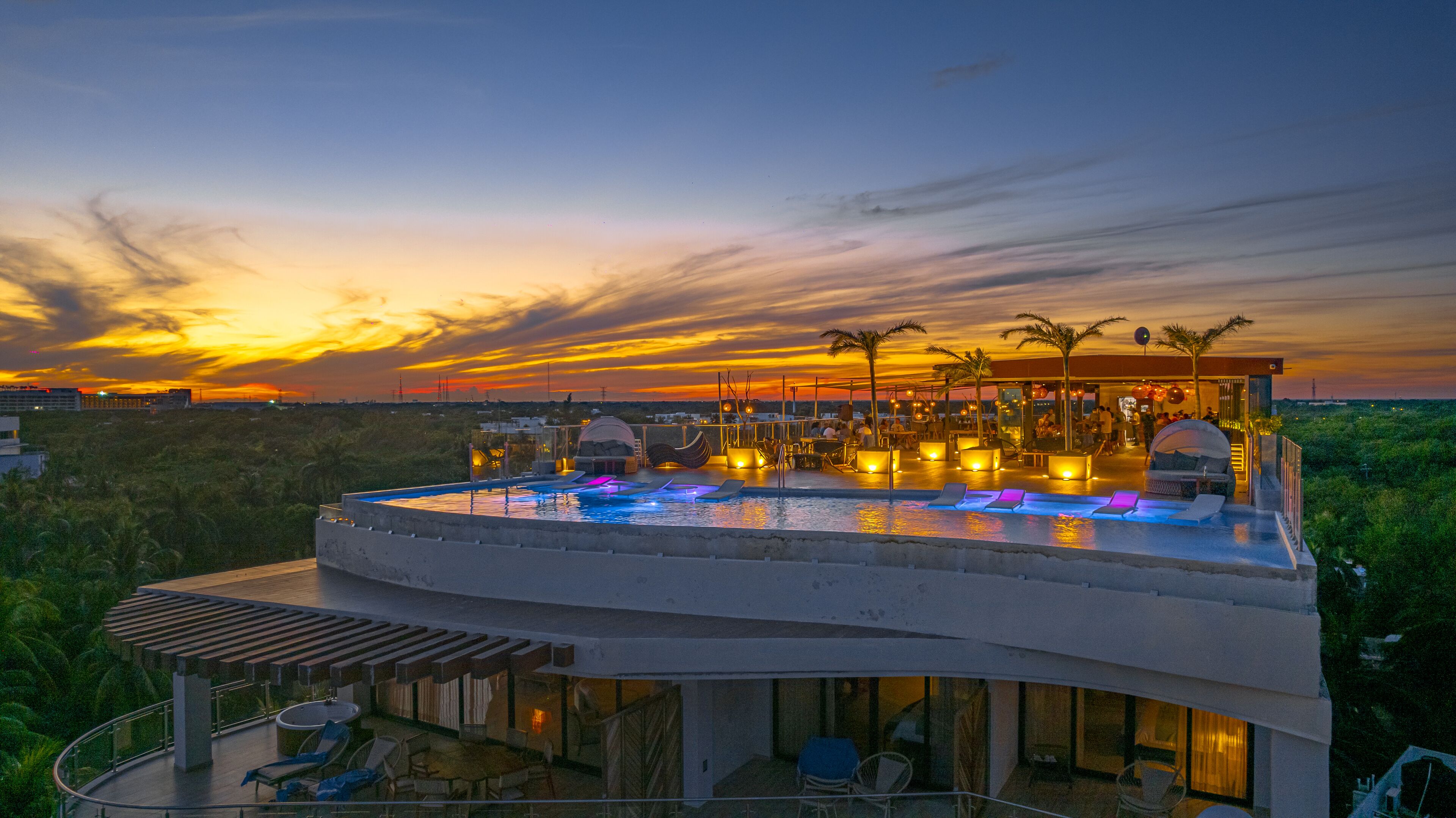 A rooftop infinity pool with lounge chairs, a bar area, and a view of the sunset.