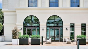 A building with a light-colored facade, large arched windows, and outdoor seating area with planters and tables.