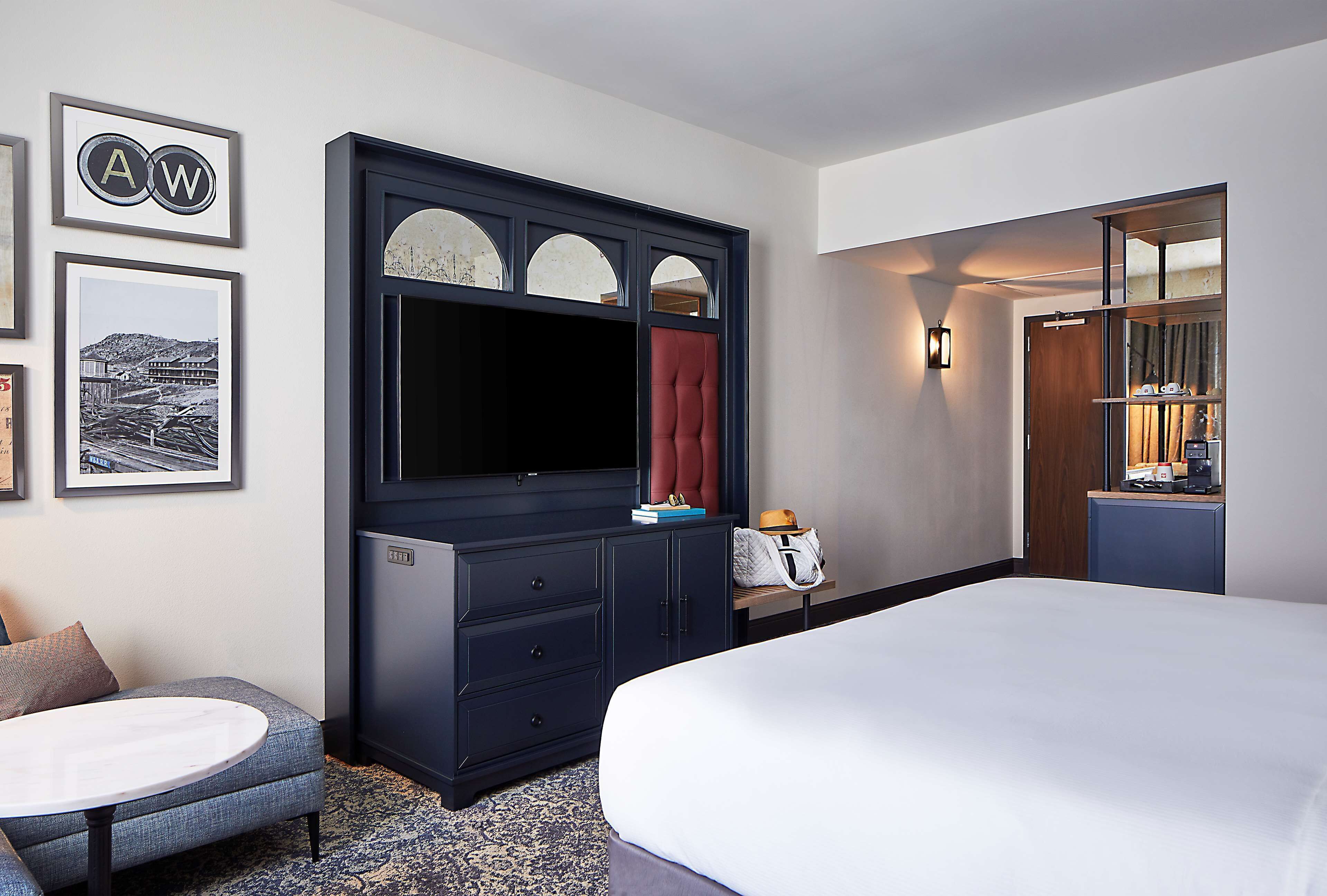 Una habitación de hotel con una cama grande, un aparador azul oscuro con un televisor de pantalla plana, una mesita redonda y un cuadro en la pared.
