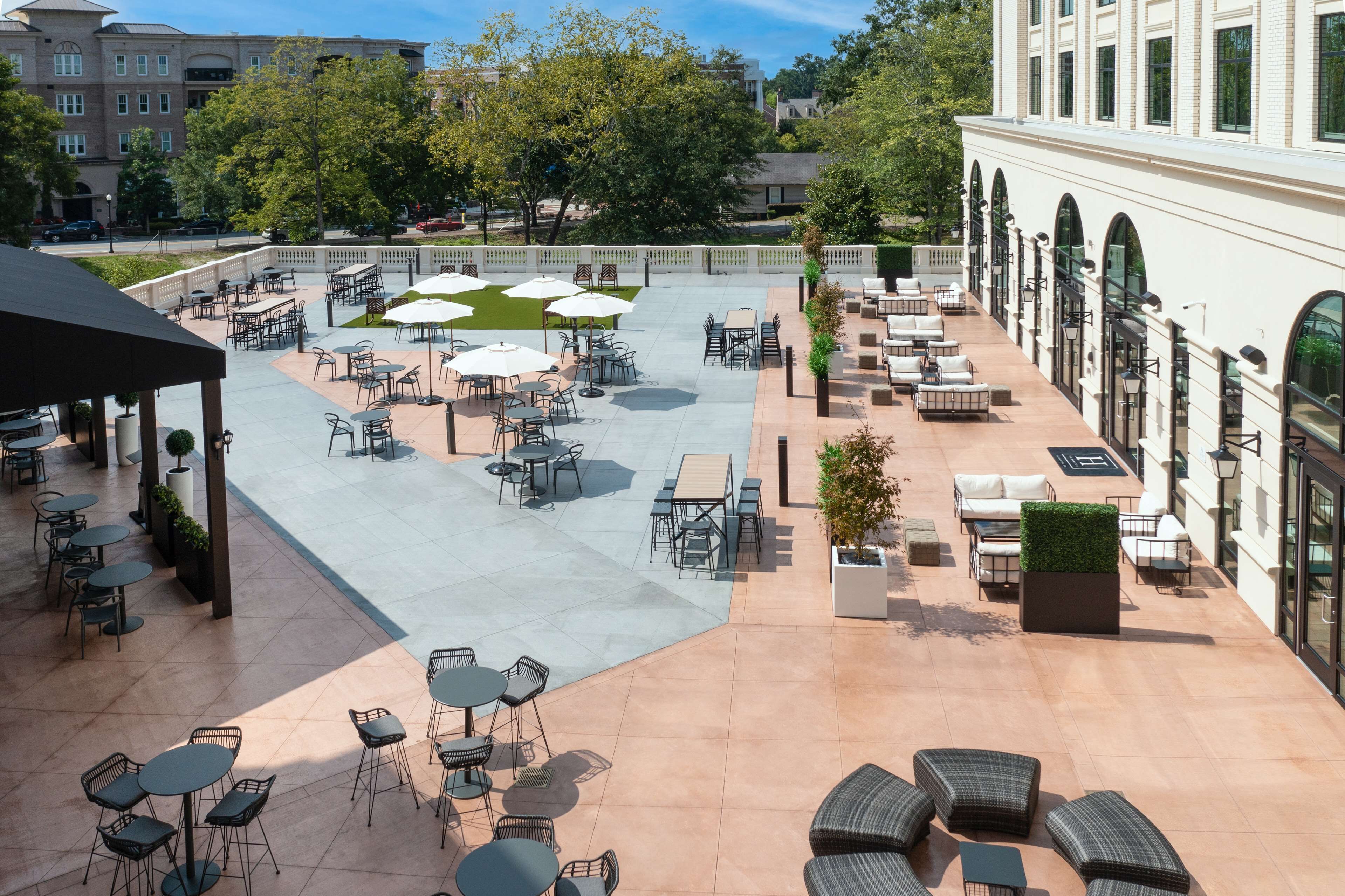A spacious outdoor patio with multiple seating areas, including tables and chairs, surrounded by greenery and a building with arched windows.