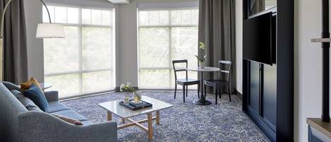 Una sala moderna con sofá, mesa de centro y televisor. Cuenta con ventanas amplias con cortinas, una mesa de comedor con sillas y una lámpara de pie.