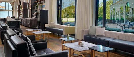 A spacious lounge area with leather sofas, wooden tables, and large windows offering views of the outdoor seating area.