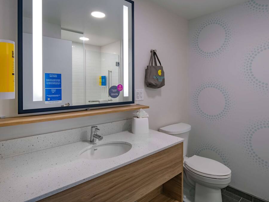 Un baño con un lavabo blanco, tocador de madera y un inodoro. Un espejo refleja la habitación, y una bolsa de tela cuelga en la pared.