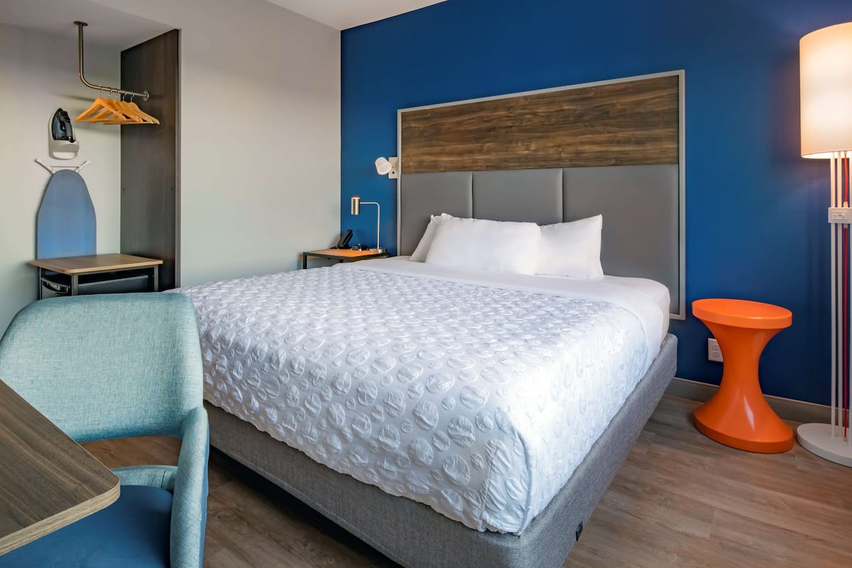 A hotel room with a bed, a desk, a chair, a nightstand, a lamp, and a wall-mounted heater.