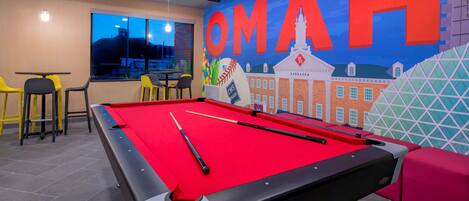 A pool table with a red felt surface, a pool cue, and a billiard ball. There is a mural on the wall with the text "OMAHA" and a depiction of a building.