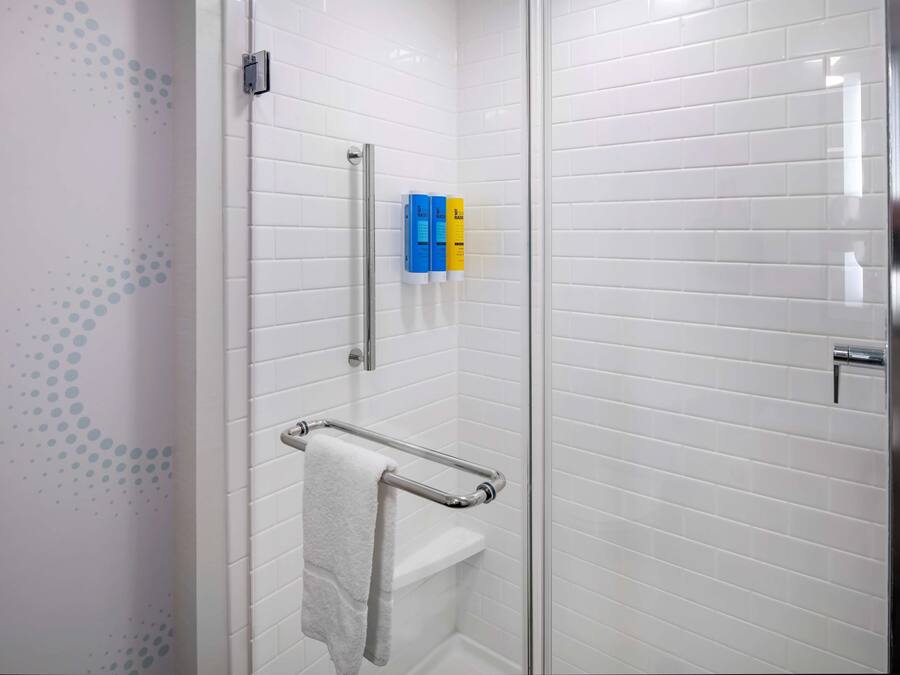 Un baño con paredes de azulejos blancos, cabina de ducha de vidrio, toallero y dos dispensadores de jabón.