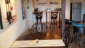 A dining area with a wooden table, chairs, and a lace table runner. There is a kitchen in the background with a refrigerator, microwave, and a coffee machine. - Gipfel Suite - Himmel Haus Bed & Breakfast (Hermann)