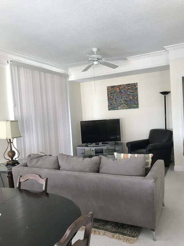Un salon avec un canapé gris, un fauteuil noir, un téléviseur à écran plat posé sur un meuble, un ventilateur de plafond et un tableau accroché au mur.