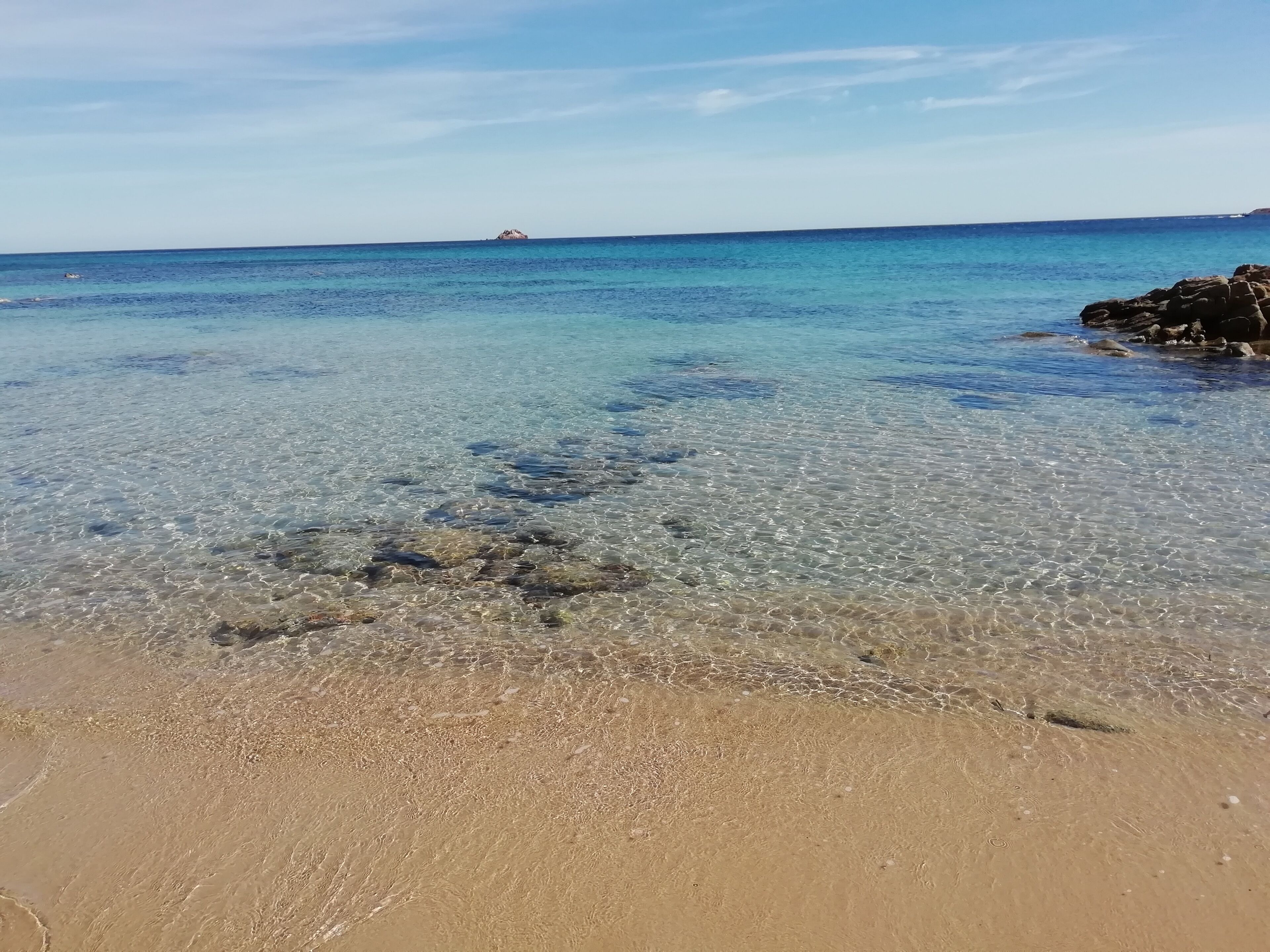 Una playa con aguas transparentes y poco profundas, un afloramiento rocoso a lo lejos y una orilla arenosa.