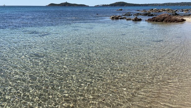 Une plage côtière aux eaux claires, bordée d’un rivage rocheux et surplombée de collines lointaines.