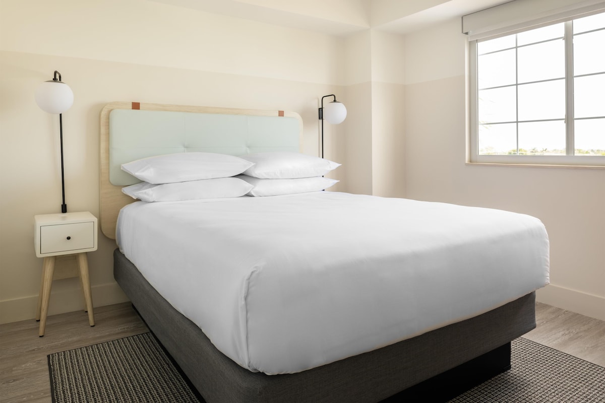 A neatly made bed with white linens and pillows, a bedside table with a lamp, and a window with a view.