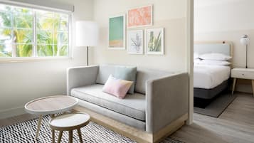 A modern bedroom with a grey sofa, a small round table, and a bed with white bedding. There are framed pictures on the wall and a window with a view of palm trees.