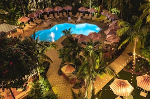 Zona de piscina con sillones de descanso y sombrillas, rodeada de palmeras y vegetación.