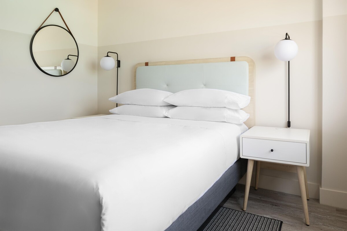 A neatly made bed with white linens, a bedside table with a lamp, and a wall-mounted mirror.