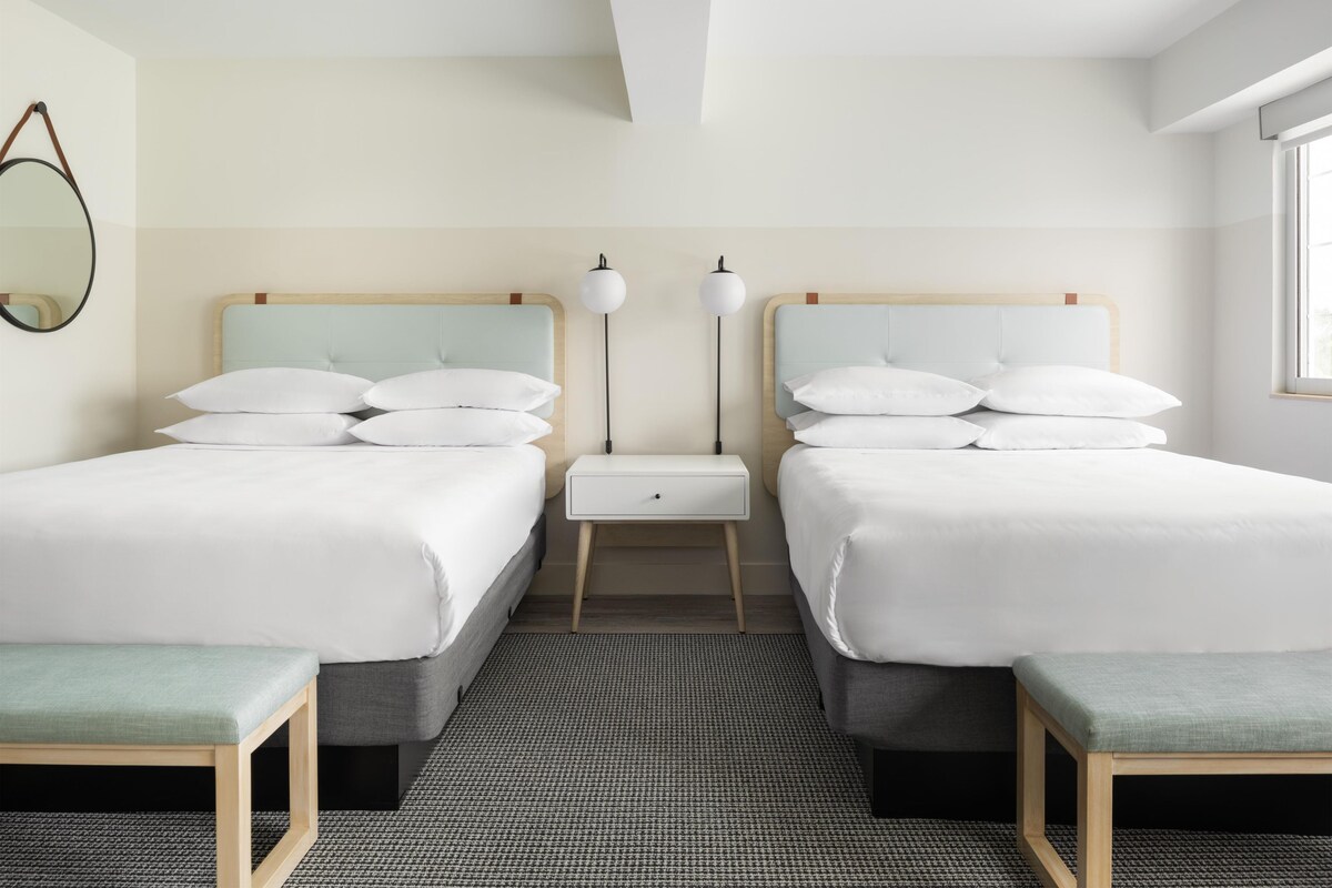 A hotel room with two beds, a bedside table, a floor lamp, and a mirror on the wall.