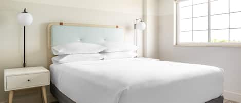 Una cama bien hecha con ropa blanca, una mesita de noche con lámpara y una ventana con persianas.