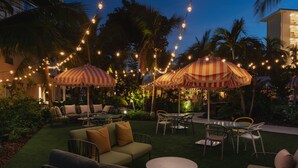 An outdoor seating area with striped umbrellas, tables, and chairs, surrounded by palm trees and string lights.