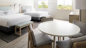 A hotel room with two beds, a round dining table, and a view of greenery through the windows.