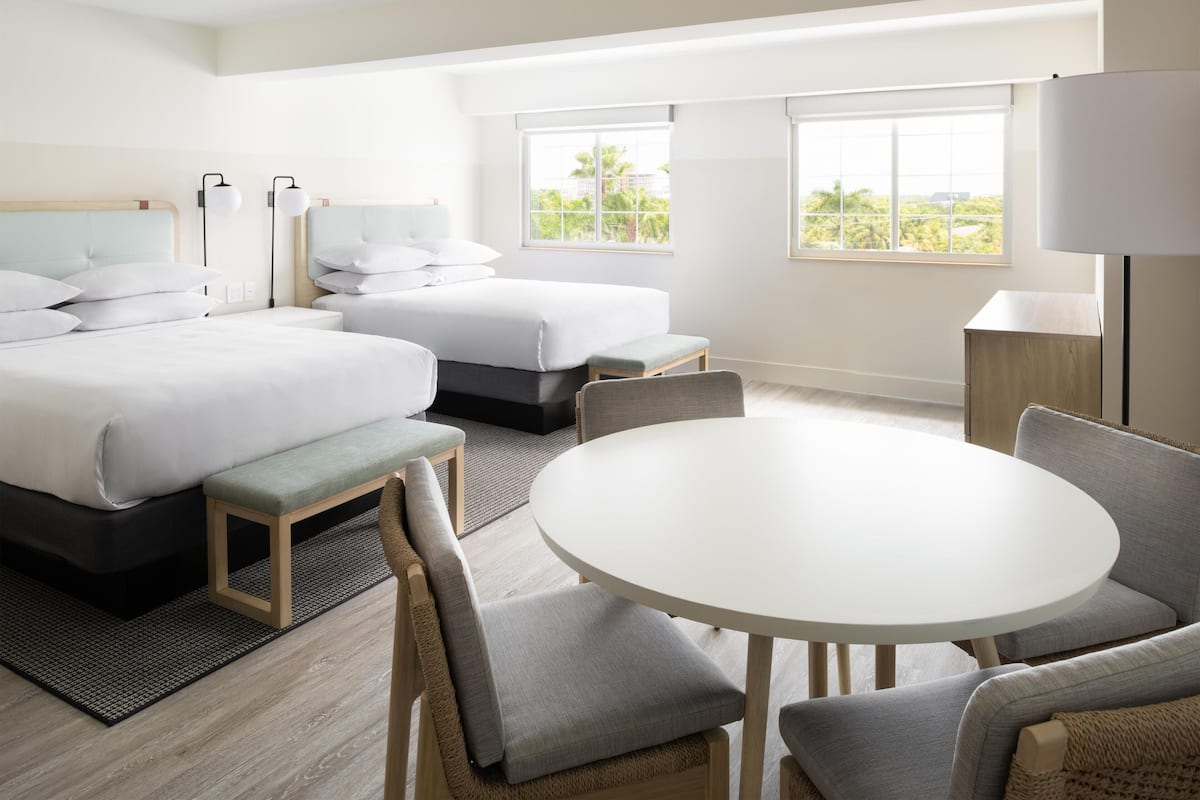 A hotel room with two beds, a round dining table, and a view of greenery through the windows.