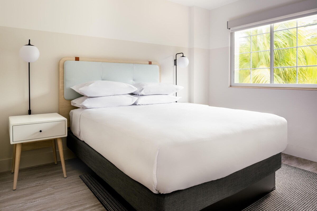 A neatly made bed with white linens, a bedside table with a lamp, and a window with a view of greenery.