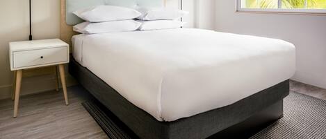 A neatly made bed with white linens, a bedside table with a lamp, and a window with a view of greenery.