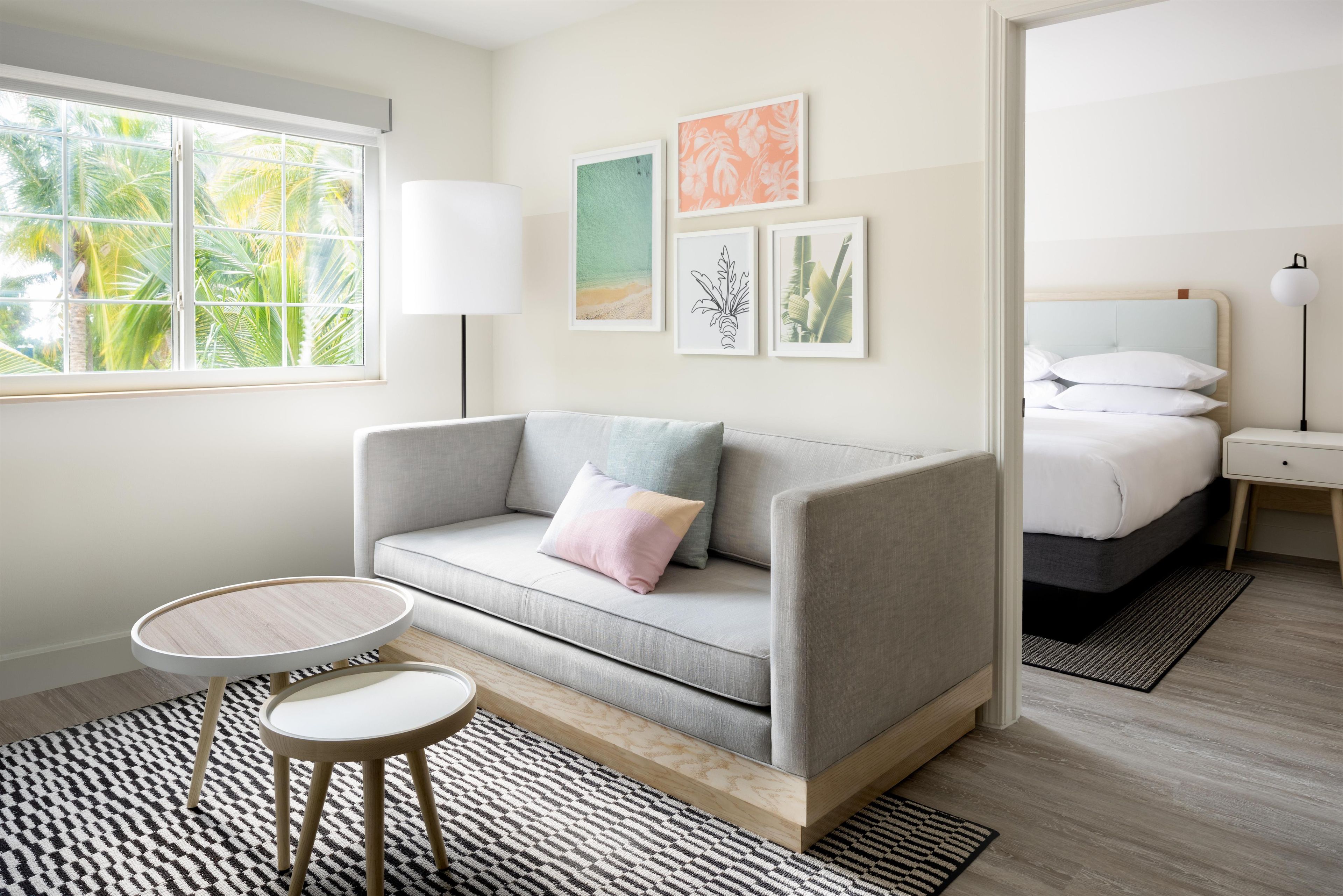 A modern bedroom with a grey sofa, a small round table, a bed with white bedding, and artwork on the wall.