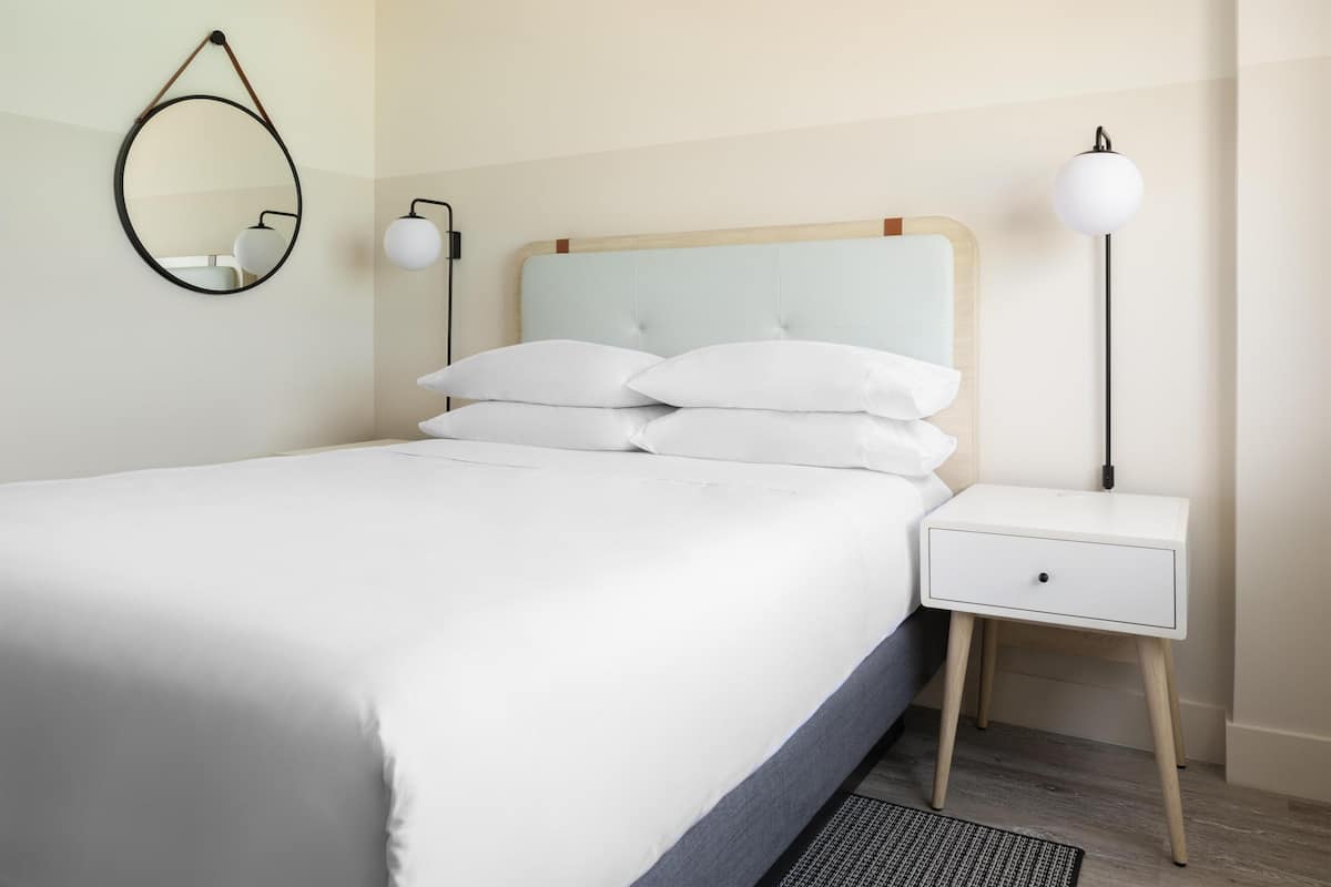 A neatly made bed with white linens, a bedside table with a lamp, and a wall-mounted mirror.