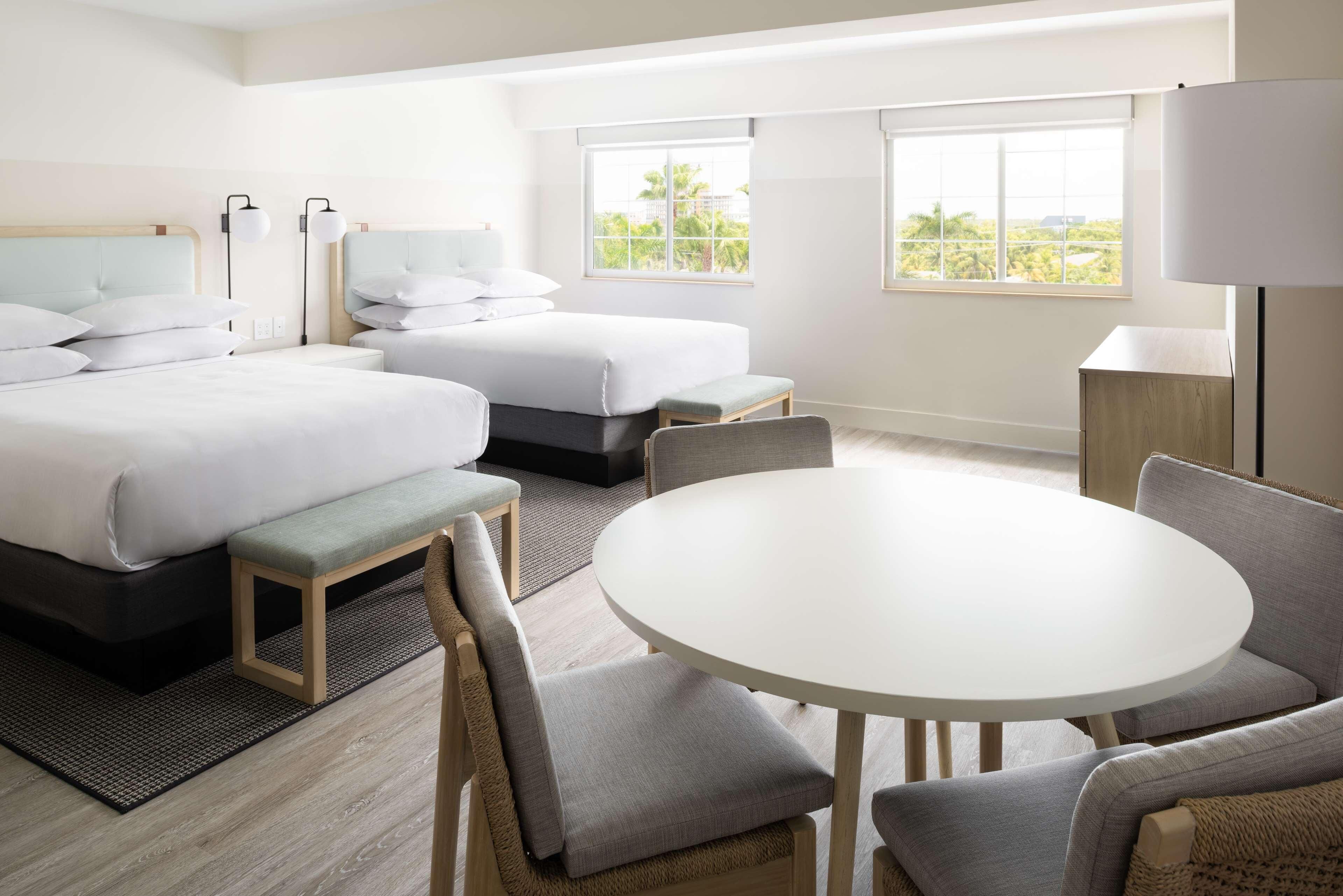 A hotel room with a bed, two bedside tables, a round dining table, and a view of greenery outside.