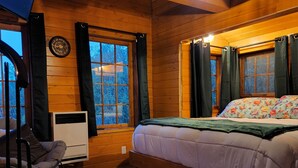 Ein gemütliches Schlafzimmer einer Holzhütte mit Holzbalkendecke, einem Bett mit Blumenmuster-Bettwäsche, einem Schaukelstuhl und einem Fenster mit Vorhängen.