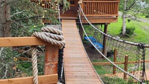 Eine hölzerne Hängebrücke mit Seilgeländern, die zu einer Holzhütte mit blauem Dach führt.