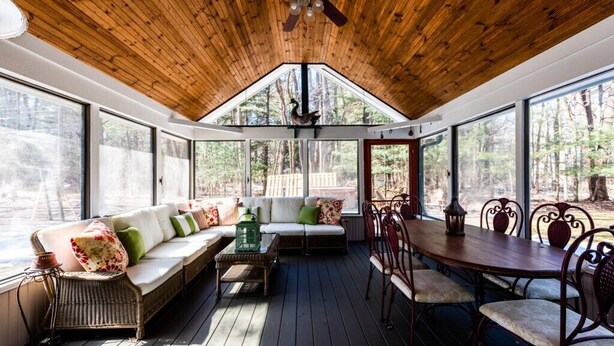A covered outdoor seating area with a wooden ceiling, large windows, a sofa with cushions, a table, and chairs. - 2027 NOW OPEN! Harbert Home With Hot Tub & Close To Beach! (Harbert)