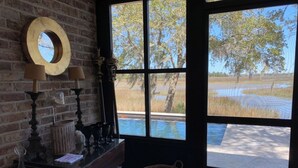 A room with a brick wall, a large window with a view of trees and water, a wooden table with a basket, and a mirror on the wall. - Water views & only 2 miles to downtown  (Charleston)