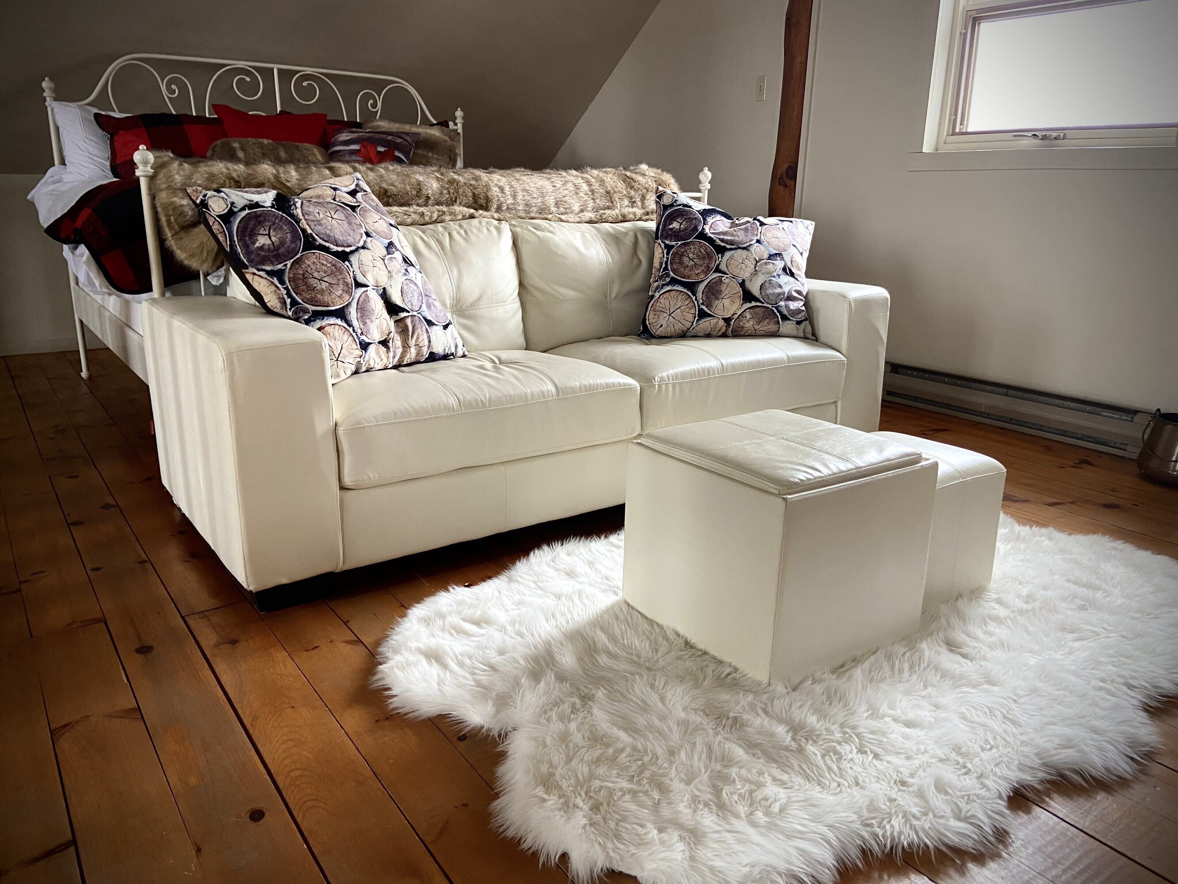 A white leather sofa with patterned cushions, a matching ottoman, and a fluffy white rug in a room with wooden flooring and a sloped ceiling.