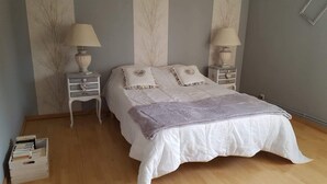 Un dormitorio con una cama, dos mesitas de noche con lámparas, una mesita auxiliar y un baúl de madera con libros.