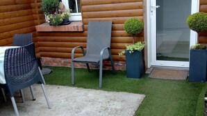 Una cabaña de troncos con un patio que tiene suelo de concreto, una silla de metal negro, una mesita y plantas en macetas.