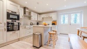 A modern kitchen with white cabinetry, a stainless steel range hood, and a built-in oven. There is a small trash bin and wooden stools near the kitchen island. - Maple House - sleeps 9 guests  in 5 bedrooms (Ebrington)