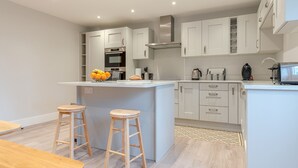 A modern kitchen with white cabinets, a central island with stools, and a built-in oven. - Maple House - sleeps 9 guests  in 5 bedrooms (Ebrington)