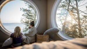 A room with two large round windows, a wicker basket, and a bed with pillows. - Waterfront Escape at the Tip of Door County (Ellison Bay)